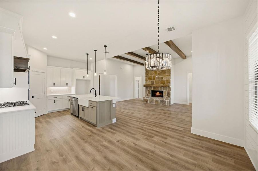 Open-concept kitchen and living area featuring wood-finish flooring, a stone-faced fireplace with a wood mantel, and exposed ceiling beams