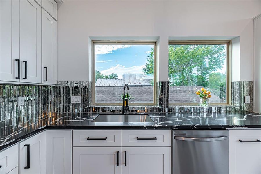 The kitchen features modern white cabinetry with black hardware, sleek black countertops, and a unique mosaic tile backsplash. Two large windows offer ample natural light and a pleasant view of the outdoors.