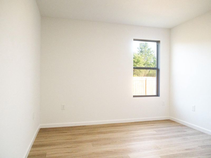 This photo shows a bright, empty room with light wood flooring and white walls. A window provides natural light and offers a view of a fence and trees outside.