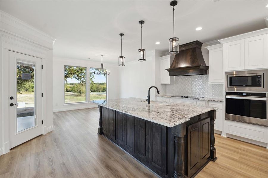 Kitchen with custom range hood, light wood-type flooring, backsplash, hanging light fixtures, and appliances with stainless steel finishes