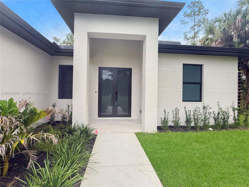 Modern stucco exterior featuring a covered entry, dark-framed double entry doors, dark-framed windows, a concrete walkway, and landscaped garden beds