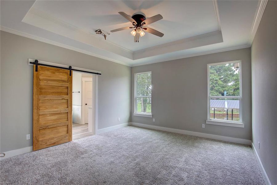 Unfurnished bedroom featuring carpet, a barn door, ceiling fan, a raised ceiling, and ornamental molding