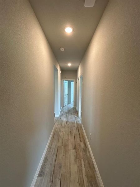 Interior hallway featuring wood-finish flooring, light neutral wall paint, recessed ceiling lighting, and white baseboards Interior hallway featuring wood-finish flooring, light neutral wall paint, recessed ceiling lighting, and white baseboards