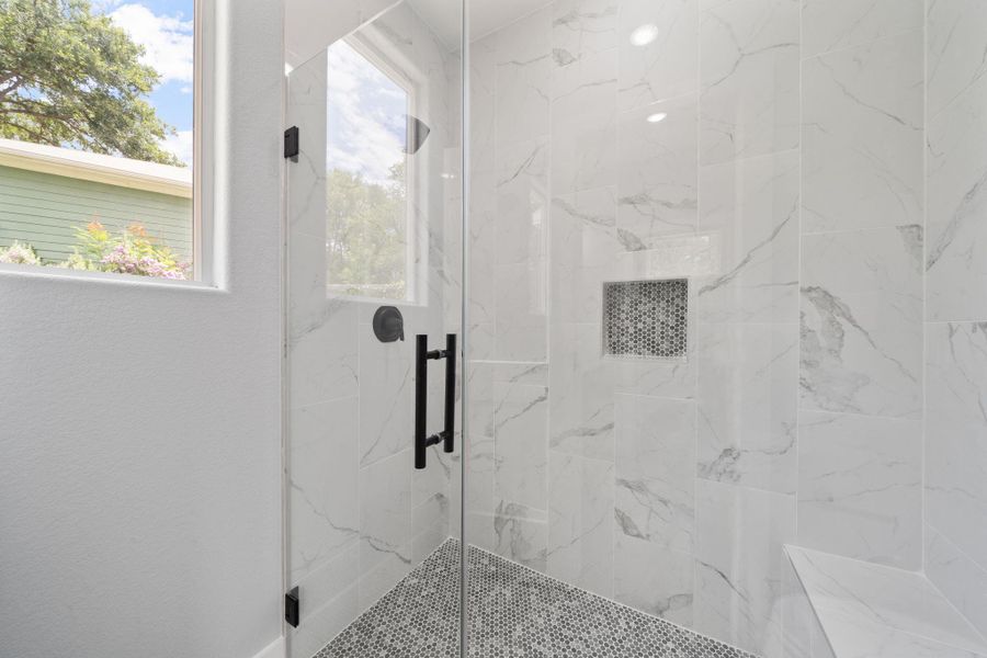 Glass-enclosed shower featuring large format white tile with grey veining, a built-in shower niche, a tiled bench, matte black fixtures, and a grey mosaic tile floor