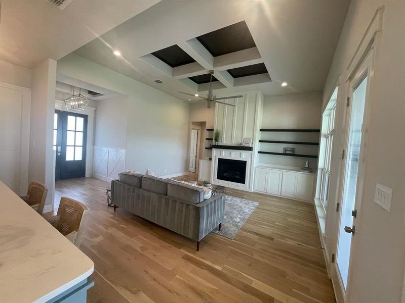 Living room with coffered ceiling, light wood-style flooring, a chandelier, a decorative wall, and a large fireplace Living room with coffered ceiling, light wood-style flooring, a chandelier, a decorative wall, and a large fireplace