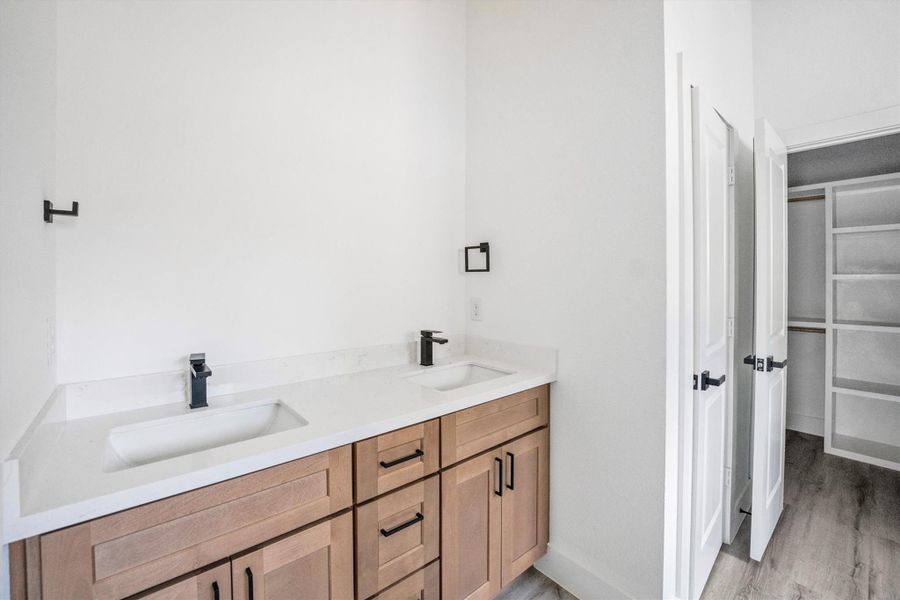 Modern bathroom with a double vanity featuring sleek black fixtures and light wood cabinets. The room includes a spacious closet with shelving and hanging space, offering ample storage.