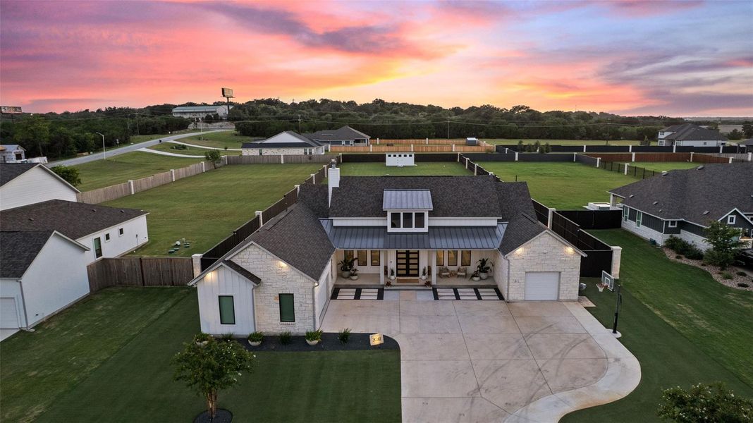 An aerial perspective reveals a modern farmhouse facade against a vibrant sunset, highlighting its spacious concrete driveway. An aerial perspective reveals a modern farmhouse facade against a vibrant sunset, highlighting its spacious concrete driveway.