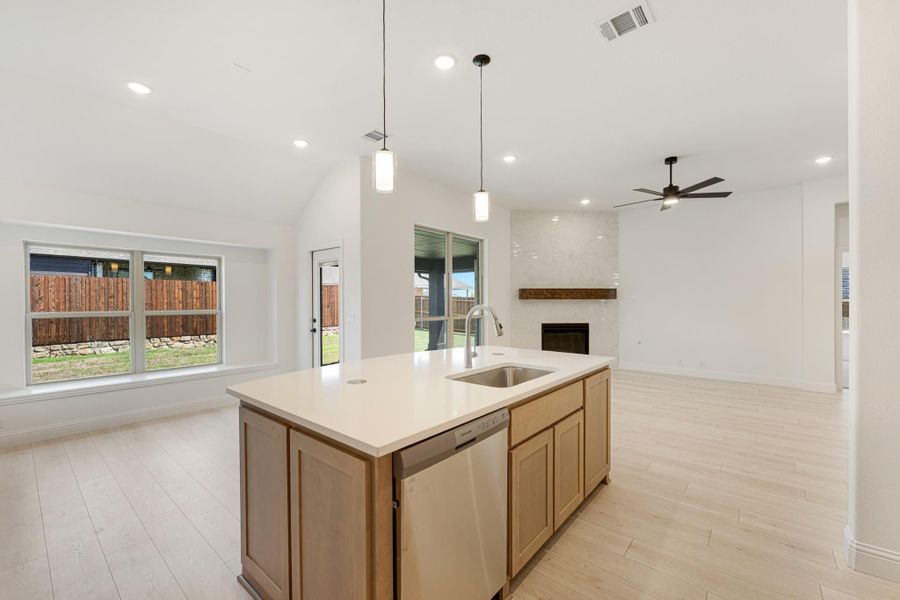 Kitchen island with sink, wood cabinets, and pendant lights, open to living room with fireplace and light wood floors