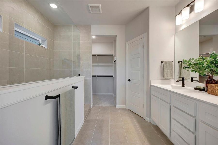 Spacious bathroom featuring a double vanity with white cabinetry and a large mirror, a walk-in shower with full-height tile surround and a transom window, and tile flooring