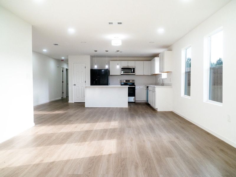 This photo showcases a bright, open-concept living area with wood-style flooring, a modern kitchen featuring white cabinetry, stainless steel appliances, and an island. Large windows provide plenty of natural light, enhancing the spacious feel.