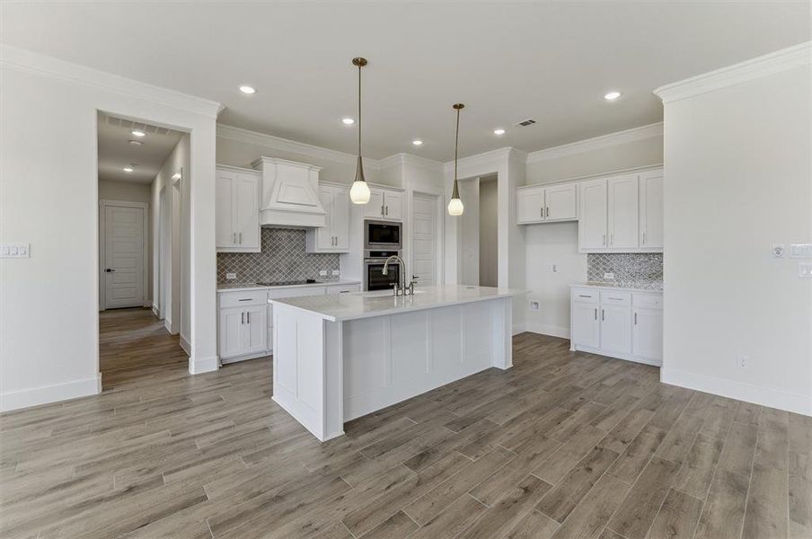 Spacious kitchen featuring an expansive central island with a sink, white cabinetry with brushed nickel hardware, integrated double ovens, and a decorative range hood