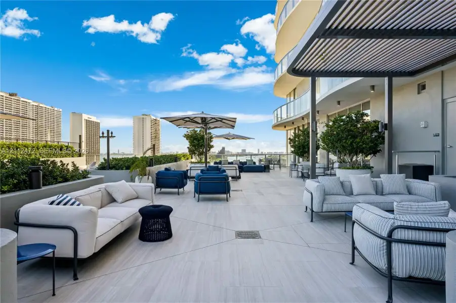 Expansive stone-tiled terrace featuring a metal louvered pergola, glass railings, built-in planter walls, and integrated exterior lighting