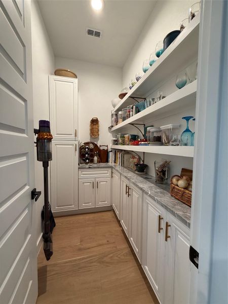 Spacious pantry featuring extensive white cabinetry, coordinating open shelving, wood-finish flooring, and gray stone countertops