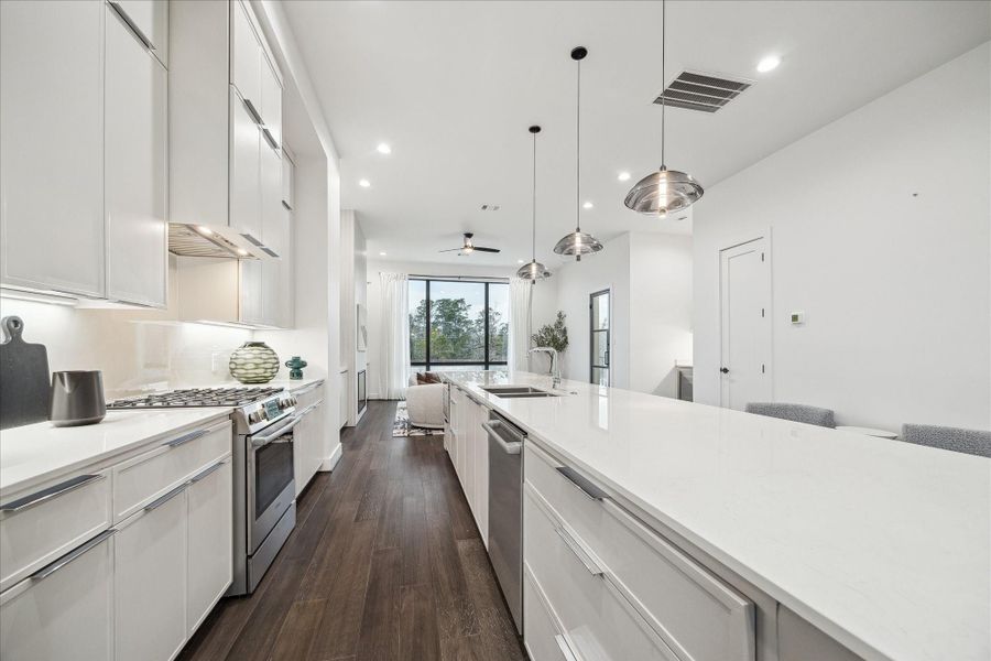 Sleek contemporary kitchen defined by a generous island with waterfall-style countertop, custom cabinetry, and under-cabinet illumination. The linear design creates clean sightlines extending toward the living area and rear windows. Elevated finishes and thoughtful layout combine to deliver both visual impact and everyday functionality in this modern culinary space.