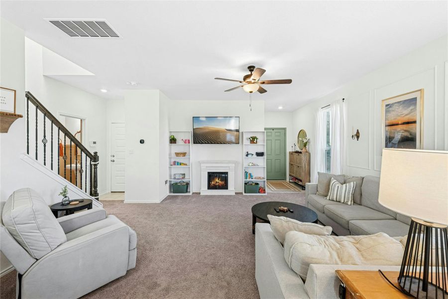 A cozy living area filled with natural light, featuring a gas fireplace flanked by decorative built-ins. This inviting space also includes carpet flooring and a ceiling fan, creating a warm and comfortable atmosphere perfect for everyday living and relaxation.