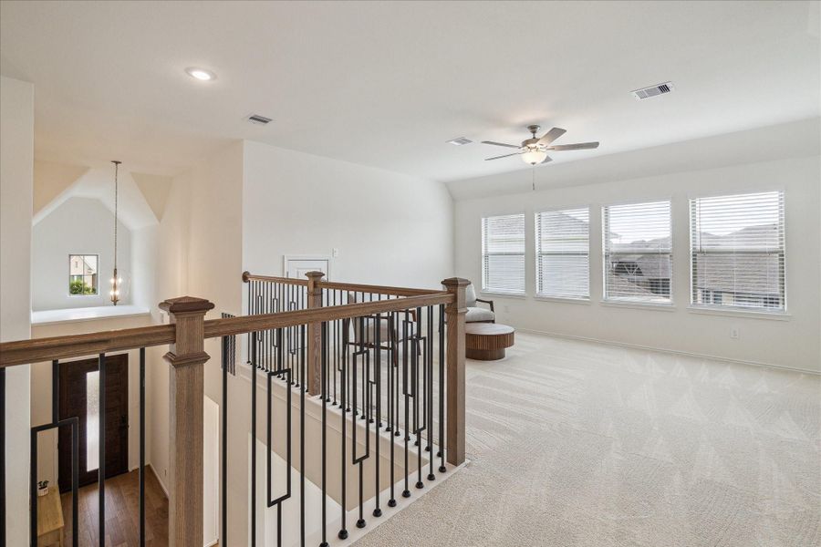 At the top of the stairs, a spacious loft/game room awaits, filled with natural light and enhanced by a ceiling fan. A sleek metal-and-wood railing provides open sightlines to the downstairs, creating a bright and connected feel.