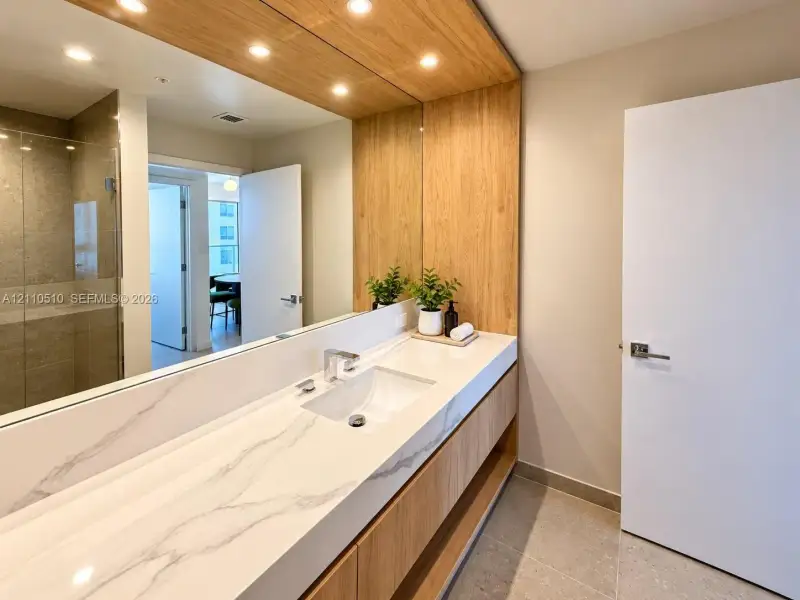 Floating vanity featuring a solid surface countertop with integrated basin, light wood cabinetry, full-width mirror with integrated wood paneling, recessed lighting, and a glass-enclosed shower with stone-finish tiling Floating vanity featuring a solid surface countertop with integrated basin, light wood cabinetry, full-width mirror with integrated wood paneling, recessed lighting, and a glass-enclosed shower with stone-finish tiling
