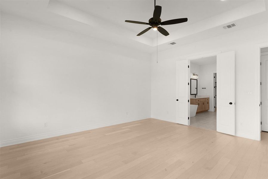 A primary bedroom with light wood flooring and a view into the primary bathroom.