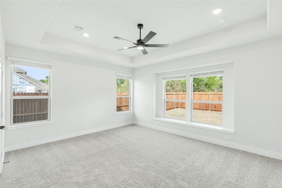 Empty bedroom with tray ceiling, ceiling fan, carpet flooring, and multiple windows overlooking backyard fence
