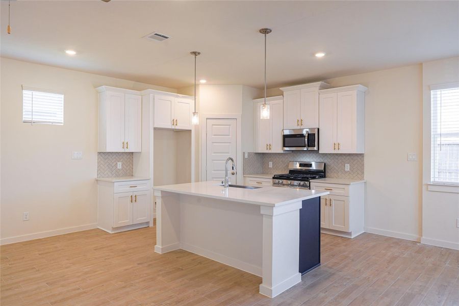 This modern kitchen features a spacious island with a sink, white cabinets, pendant lighting, and stainless steel appliances. The light wood flooring and neutral walls create a bright, inviting space.
