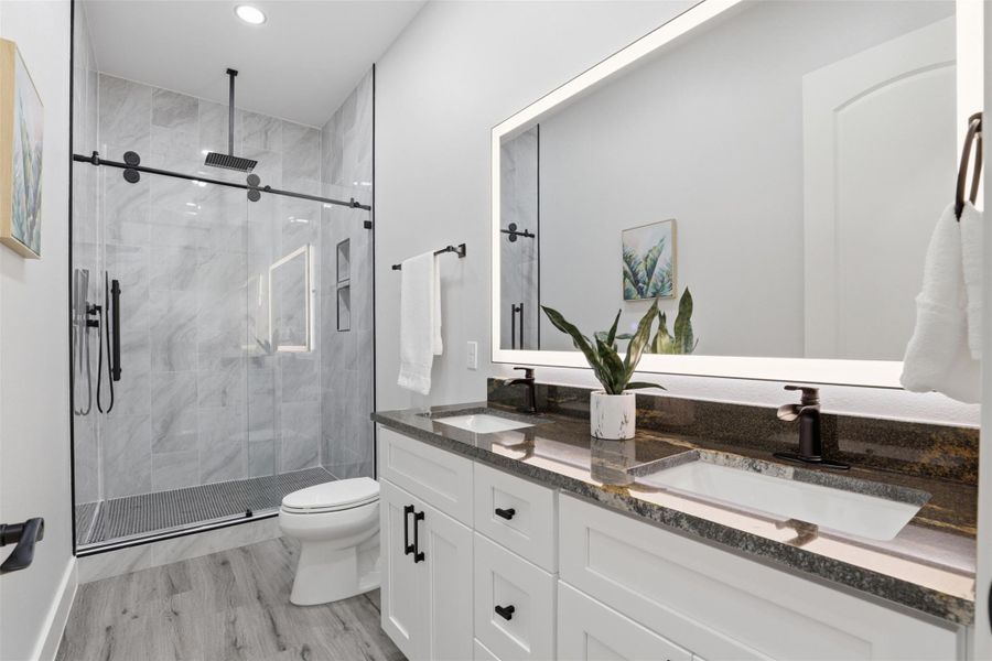 Contemporary bathroom featuring a dual vanity with dark countertops and undermount sinks, an illuminated mirror, a glass-enclosed shower with a rain shower head and matte black fixtures, and wood-finish flooring
