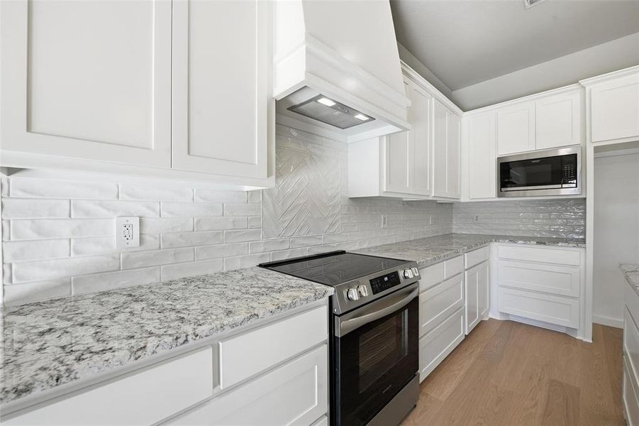 Kitchen with appliances with stainless steel finishes, white cabinetry, light stone counters, and custom range hood Kitchen with appliances with stainless steel finishes, white cabinetry, light stone counters, and custom range hood