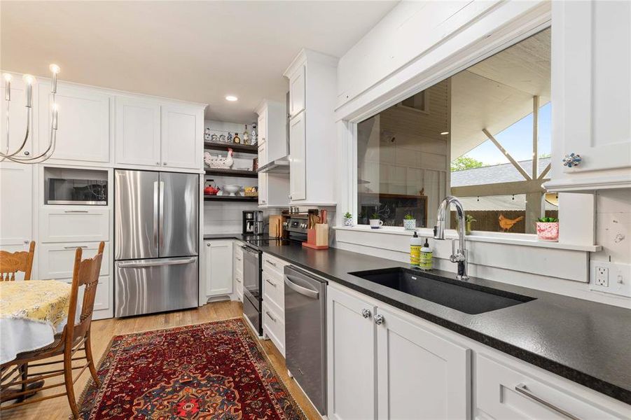 Modern kitchen featuring white shaker cabinetry, stainless steel appliances, dark countertops, and a large window overlooking an outdoor covered patio