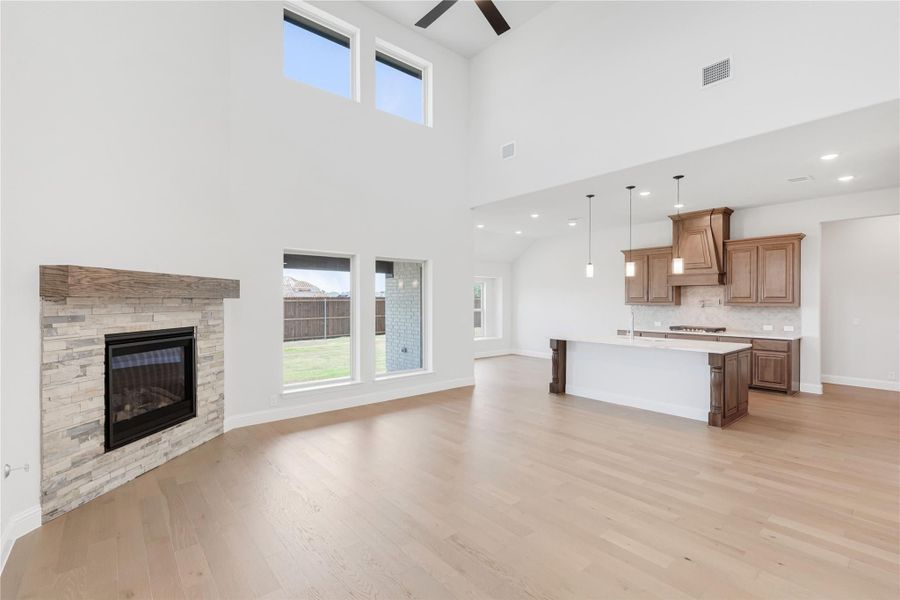 Open-concept living room with stone fireplace, high ceilings, and hardwood floors open to kitchen with wood cabinets Open-concept living room with stone fireplace, high ceilings, and hardwood floors open to kitchen with wood cabinets