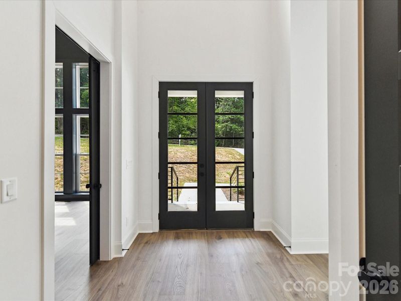 Bright, welcoming entry hall with wide plank flooring, modern black doors, and an open sightline to the light-filled living area with views of the outdoors.