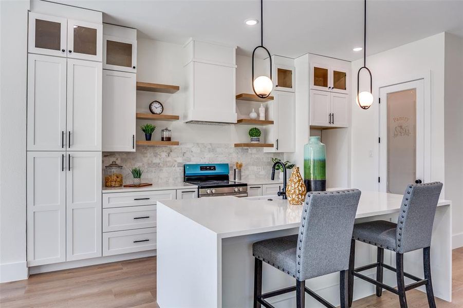 Kitchen with stainless steel range with electric stovetop, open shelves, light countertops, a kitchen island with sink, and white cabinets Kitchen with stainless steel range with electric stovetop, open shelves, light countertops, a kitchen island with sink, and white cabinets