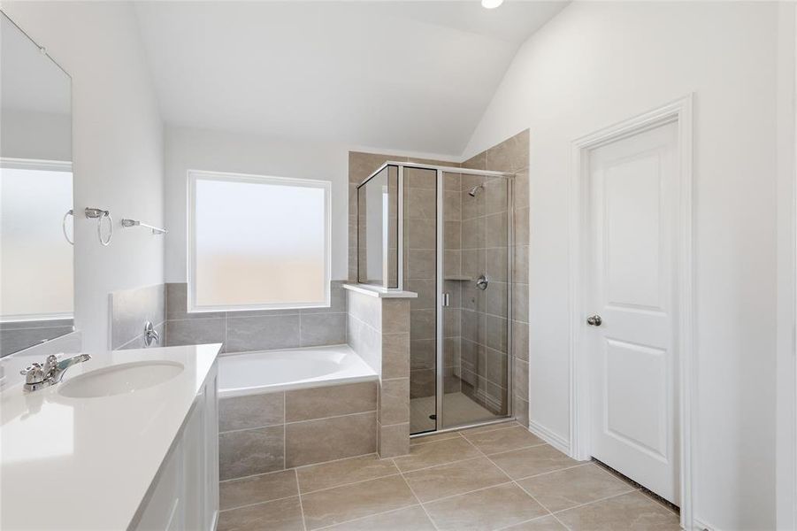 Bathroom featuring a large soaking tub with tiled surround, a separate glass-enclosed shower, a vanity with a white countertop and integrated sink, a large wall mirror, and neutral-toned floor tiling