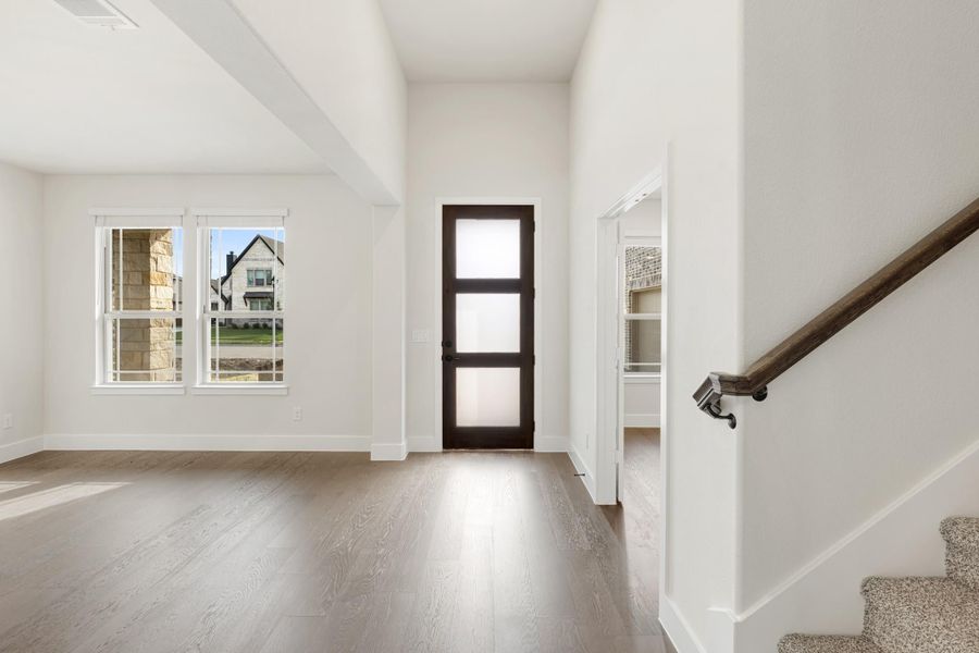 Open entryway with hardwood floors, dark glass-panel door, white walls, and staircase with wood handrail