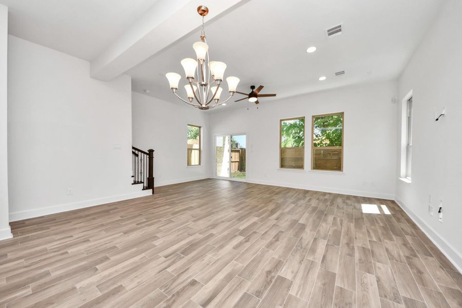 This spacious living area features elegant ceramic wood-look flooring, large windows for natural light, a stylish chandelier, and a ceiling fan. The open layout leads to a staircase and a view of the backyard.