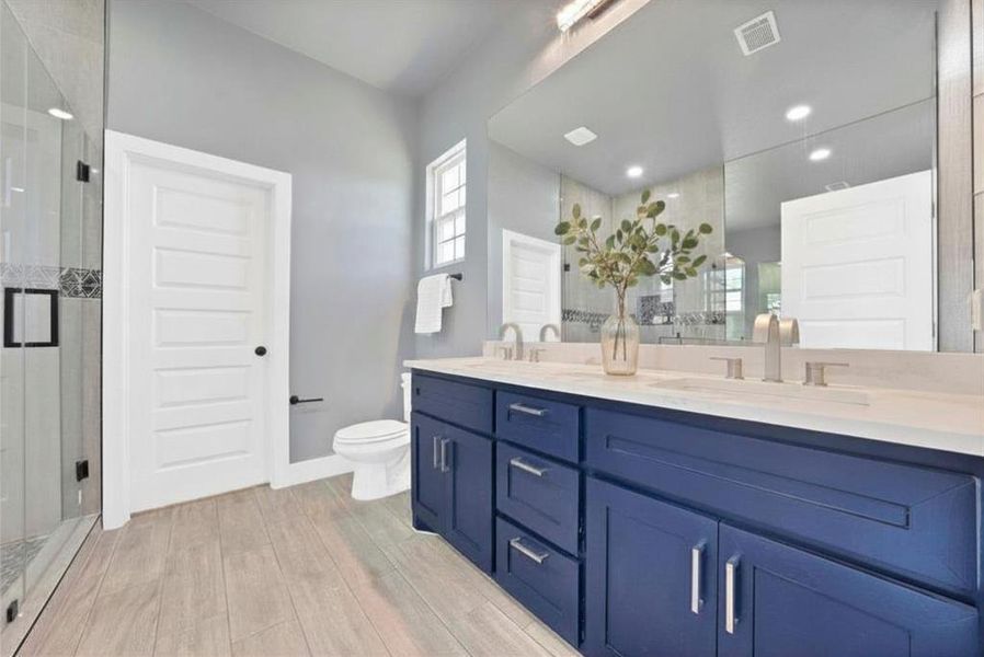 Dual vanity with a white countertop and navy cabinetry, wood-finish flooring, full-width mirror, walk-in shower with glass enclosure, and a frosted window