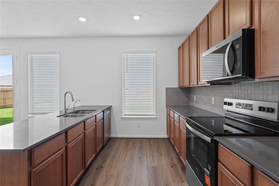 This kitchen boasts high ceilings, Woodmont Cody cabinets in Toffee stain, Grey Expo silestone countertops, stainless steel Whirlpool appliances, a modern Daltile backsplash, recessed lighting, and a pantry—all overlooking the expansive Great Room.