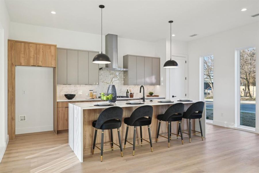 Kitchen featuring an island with sink, decorative backsplash, hanging light fixtures, wall chimney exhaust hood, and light hardwood / wood-style flooring