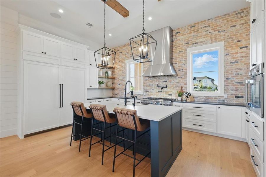 Kitchen with wall chimney exhaust hood, stainless steel oven, light wood-style flooring, an island with sink, and hanging light fixtures Kitchen with wall chimney exhaust hood, stainless steel oven, light wood-style flooring, an island with sink, and hanging light fixtures