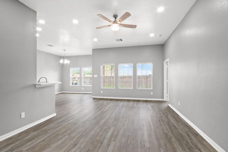 Bright and spacious open-concept living area with modern gray walls and wood flooring. Features a ceiling fan, large windows for natural light, and a view into the kitchen with a breakfast bar. Ideal for comfortable living and entertaining.