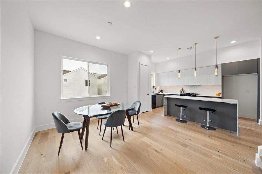 Bright and modern open-concept kitchen and dining area with light wood flooring, sleek cabinetry, and pendant lighting. Features a spacious island with seating and a large window for natural light.