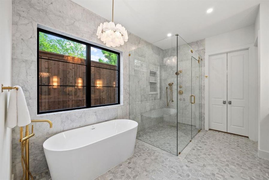 Luxurious bathroom featuring a freestanding soaking tub with a gold-tone floor-mounted faucet, a spacious glass-enclosed shower with a built-in bench, and marble-tiled walls