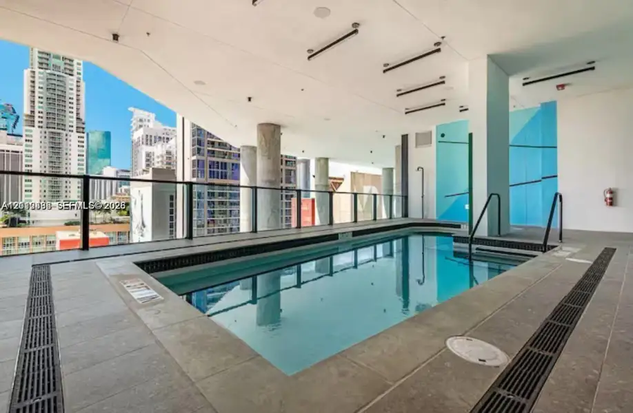 Covered swimming pool featuring large-format tile decking, structural concrete columns, a sleek glass railing, and linear ceiling lighting Covered swimming pool featuring large-format tile decking, structural concrete columns, a sleek glass railing, and linear ceiling lighting