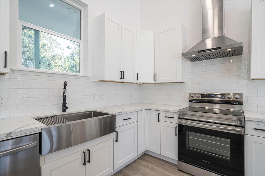 Kitchen with stainless steel appliances, wall chimney exhaust hood, light stone counters, white cabinetry, and tasteful backsplash