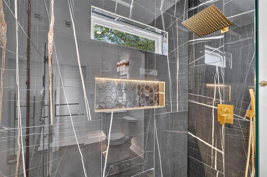 Shower enclosure featuring large format dark tile with white veining, a built-in illuminated niche with hexagonal tile, a gold-finish rain shower head, gold-finish shower controls, and a clear glass shower door