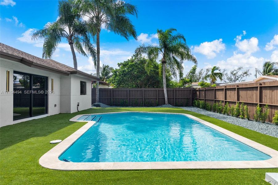 Outdoor living area featuring a rectangular swimming pool with stone coping, synthetic turf, privacy wood fencing, and mature palm trees