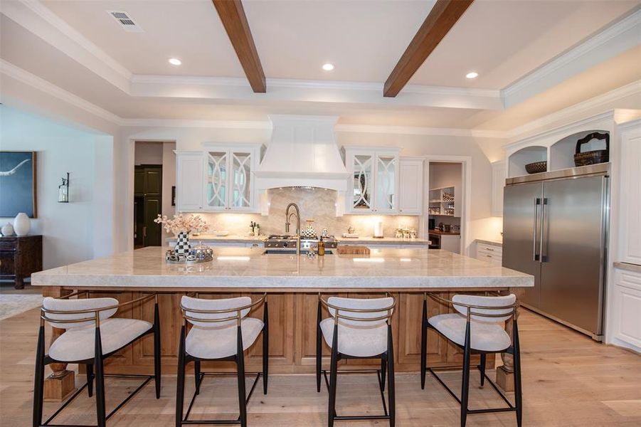 This kitchen features a spacious island with seating for four, elegant marble countertops, and modern stainless steel appliances. It has an open layout with high ceilings accented by wooden beams, ample cabinetry, and stylish lighting, creating a warm and inviting atmosphere.