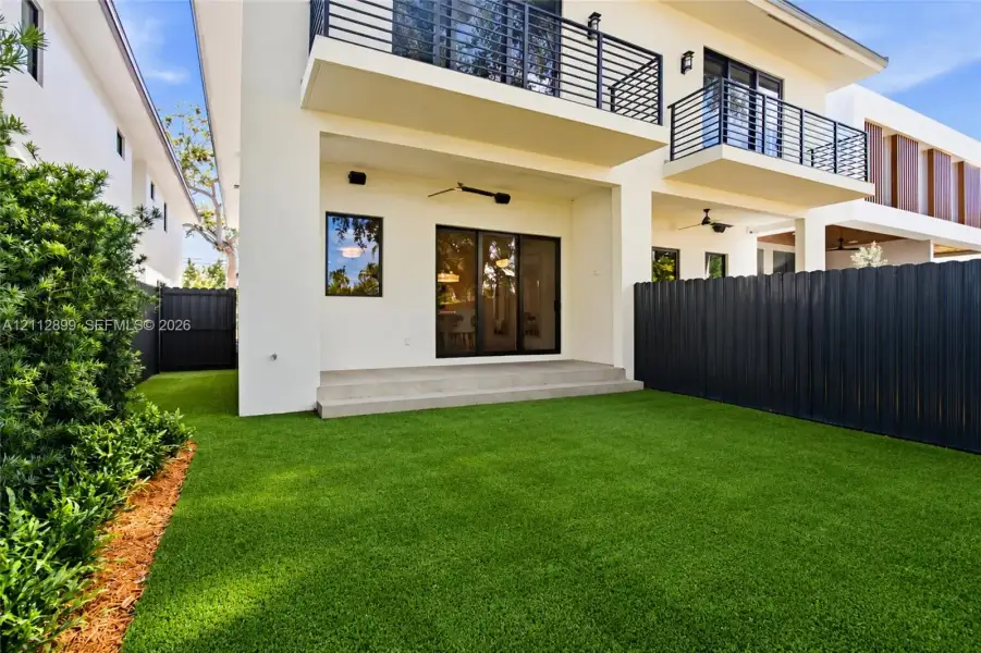 Rear exterior featuring a concrete patio, synthetic turf lawn, privacy fencing, and second-level balcony with metal railings
