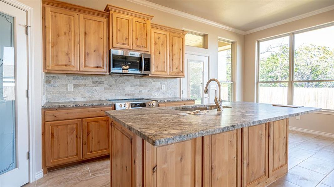 Kitchen featuring ornamental molding, backsplash, light stone counters, stainless steel appliances, and a kitchen island with sink