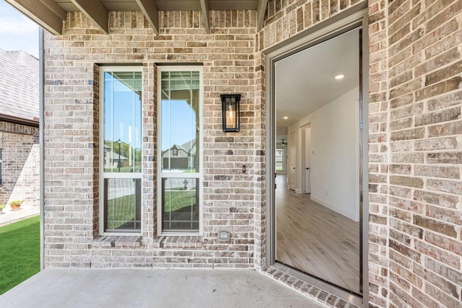 Home entry with light brick exterior, open front door revealing hardwood floors, and wall-mounted lantern light fixture Home entry with light brick exterior, open front door revealing hardwood floors, and wall-mounted lantern light fixture