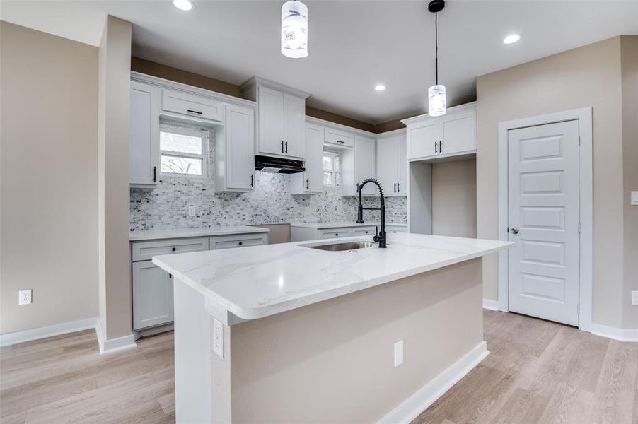 Kitchen with pendant lighting, a sink, light stone counters, under cabinet range hood, and backsplash