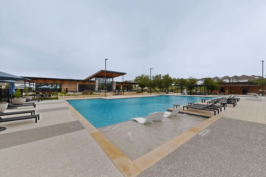 Community pool featuring a shallow water lounging area with seating, surrounding paved sundeck, a modern clubhouse with expansive windows, and shaded outdoor seating areas